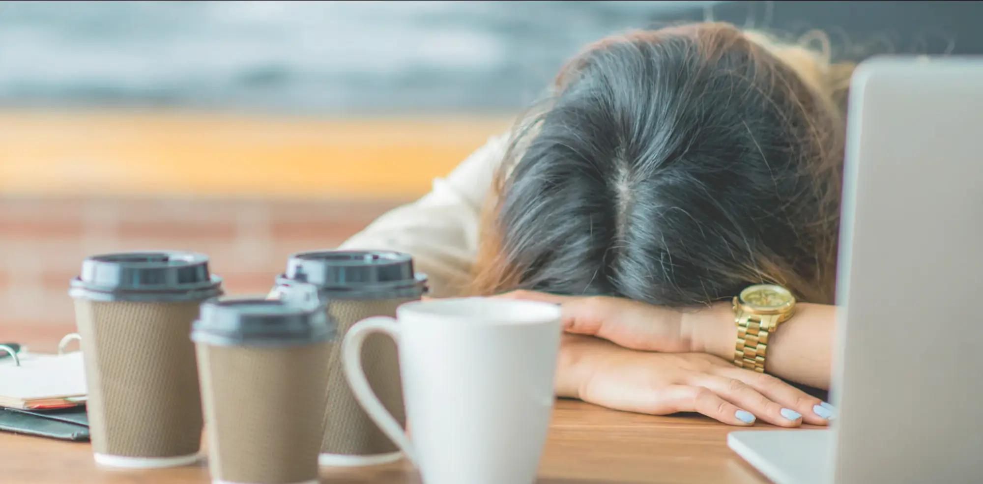 Madre con la cabeza apoyada en la mesa mostrando signos de agotamiento emocional, rodeada de café y trabajo acumulado.