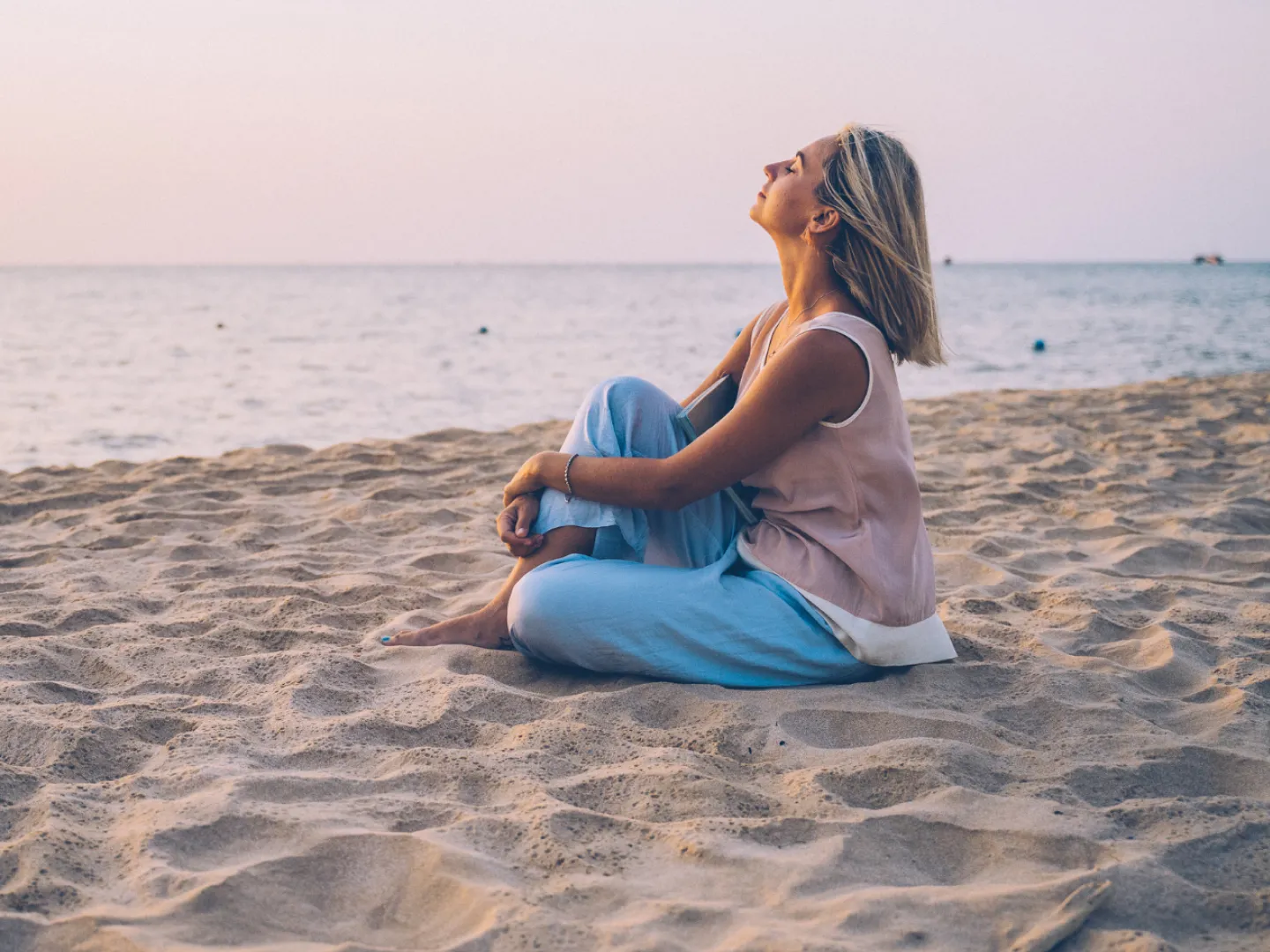 Foto de una mujer sentada en la arena de una playa, con los ojos cerrados aceptando otras formas de maternidad. logo principal Rocío Mesa. Terapéuta emocional y life coach para madres. Presencial y online en Palma de Mallorca